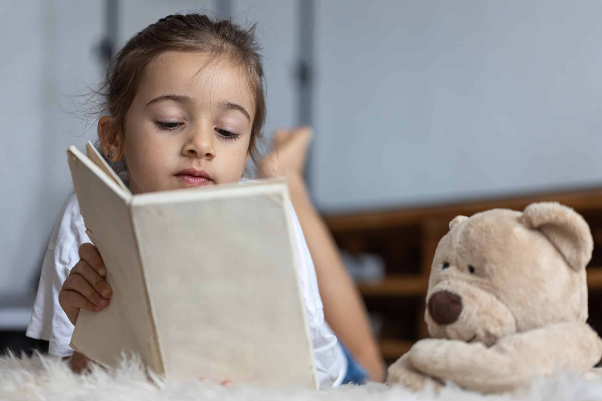 Little girl reads a book with a teddy bear.